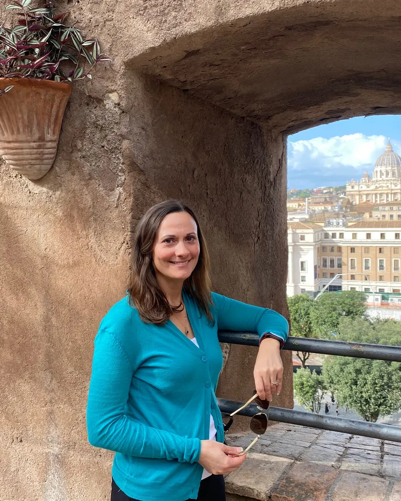 Jessica in a bright teal cardigan, framed by a stone archway with St. Peter’s Basilica in the background